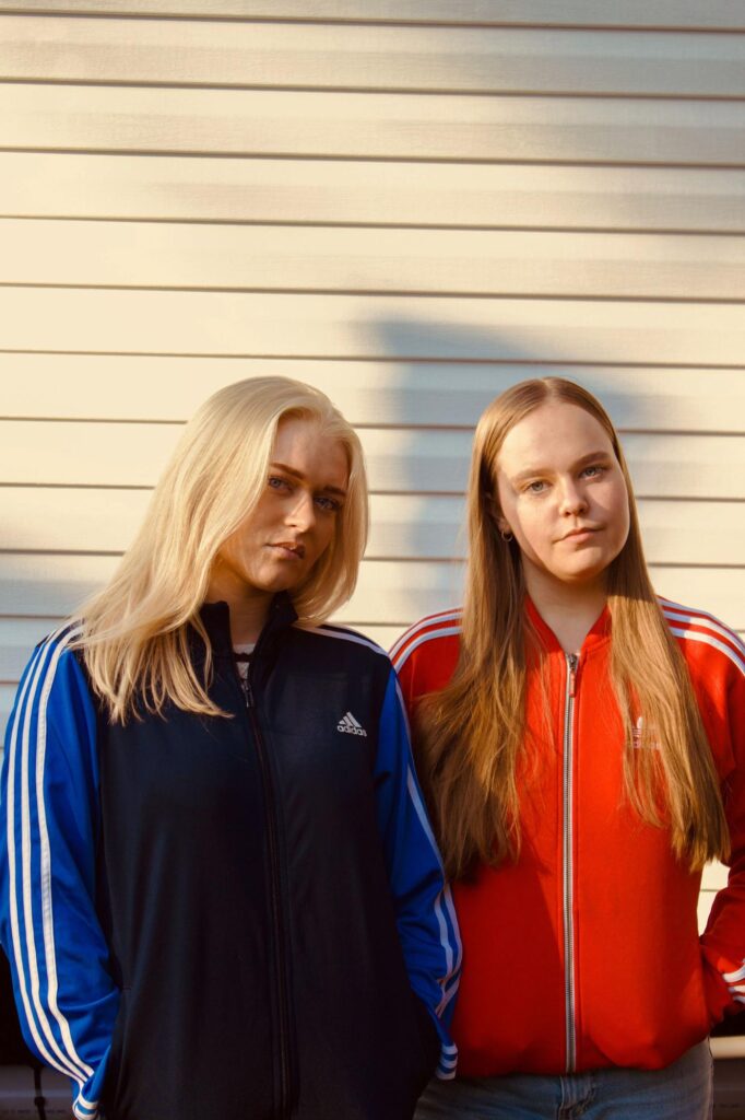 Two young women stand side by side against a white wall, both wearing sporty jackets—one in navy blue with blue and white stripes, the other in red with white stripes. They look directly at the camera with neutral expressions.