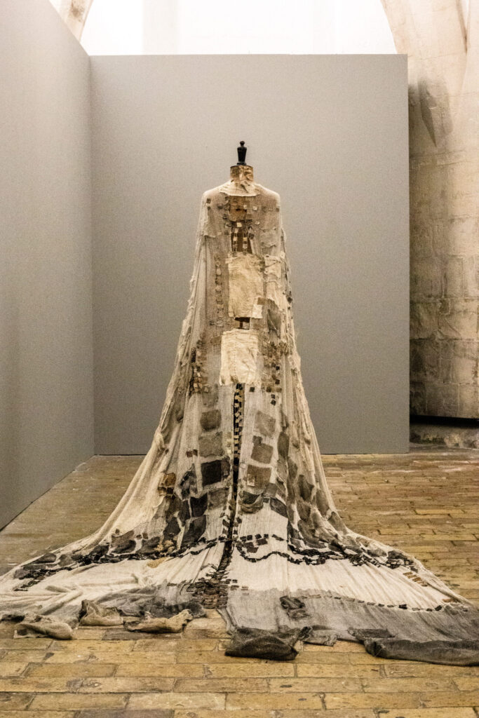 A long, ornate, textured gown with patchwork and draped fabric, displayed on a dress form in a minimalist gallery space with stone floors and neutral-colored walls.