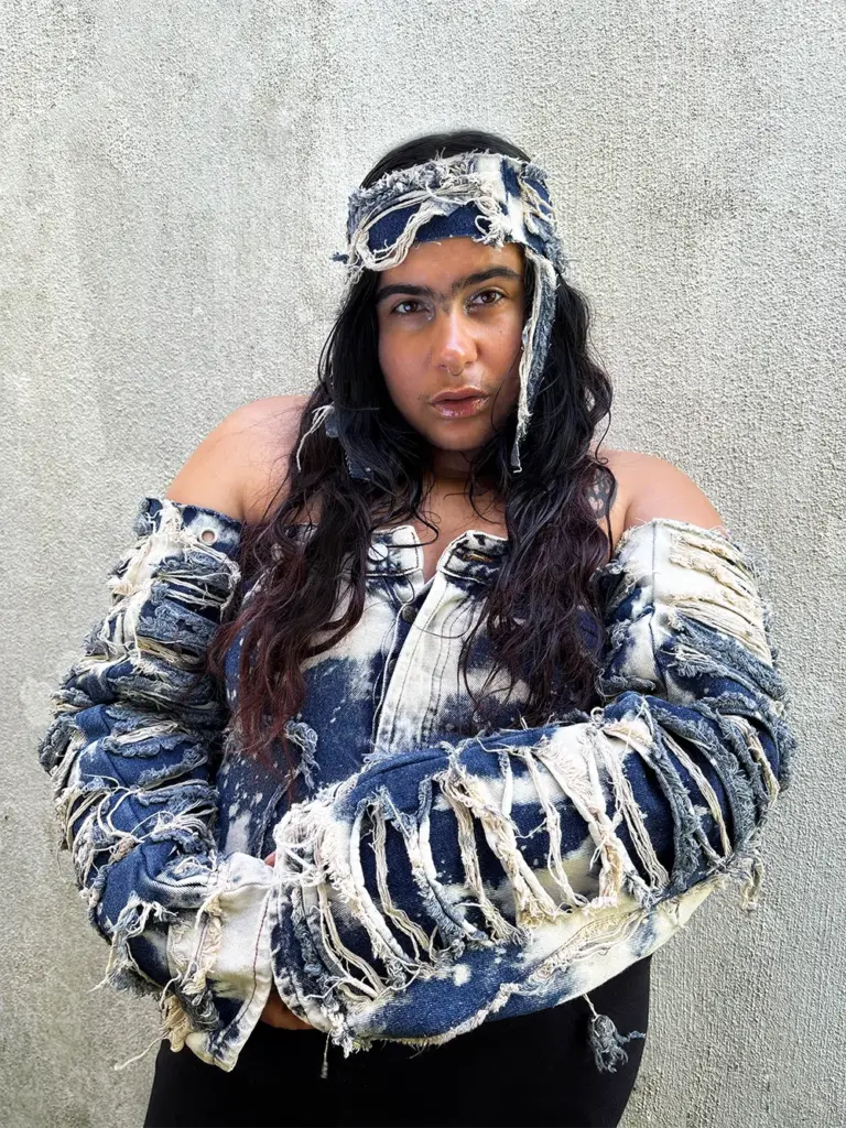 A person poses against a textured wall, wearing a distressed, ripped blue denim jacket and headband. Their long dark hair falls over their shoulders.