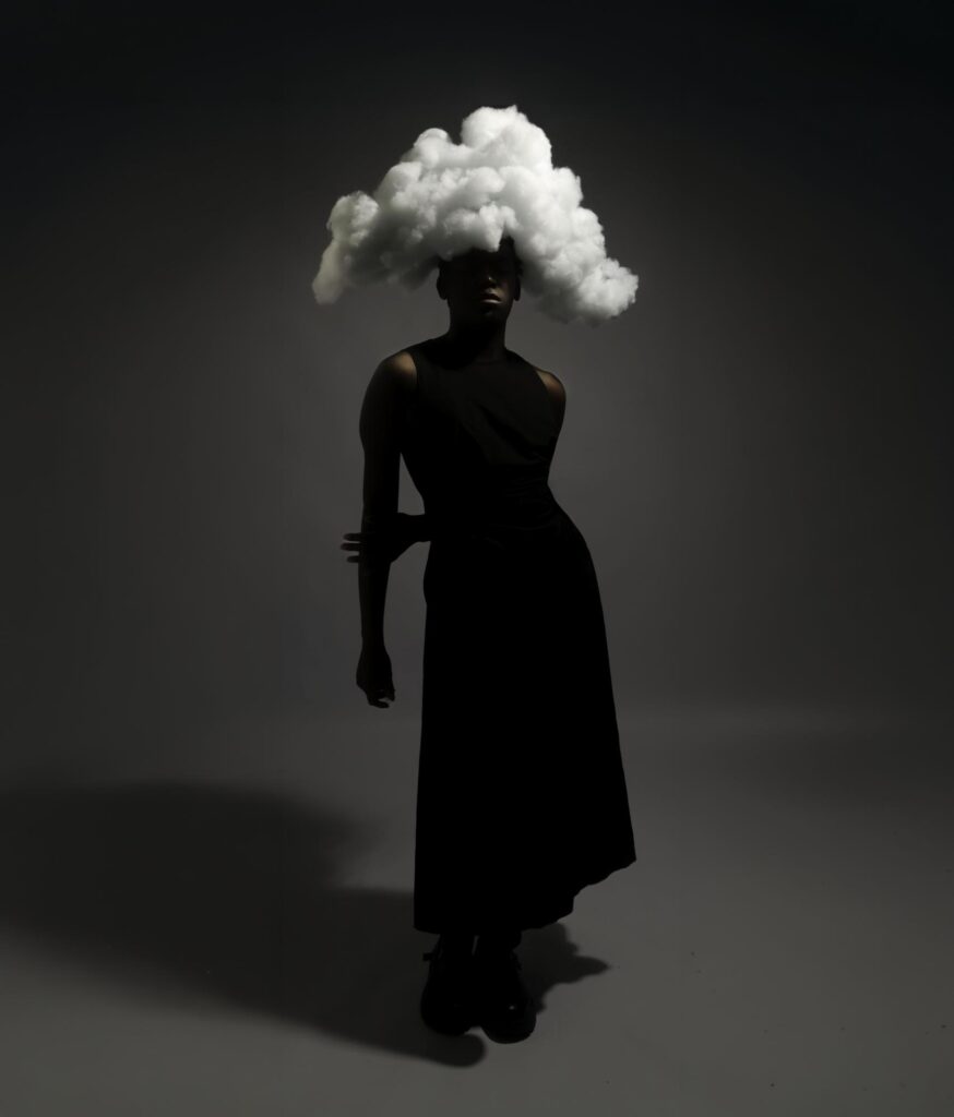 A person in a black dress stands in a dark room with a large, fluffy white cloud prop covering their head, casting dramatic shadows.