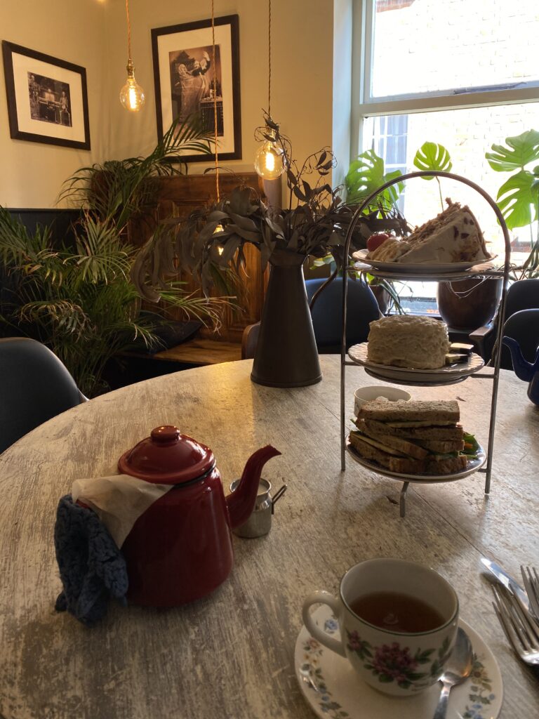 A cozy café table set for afternoon tea with a red teapot, a floral teacup, and a three-tier stand holding sandwiches and cakes. The background has plants, framed photos, and warm lighting.