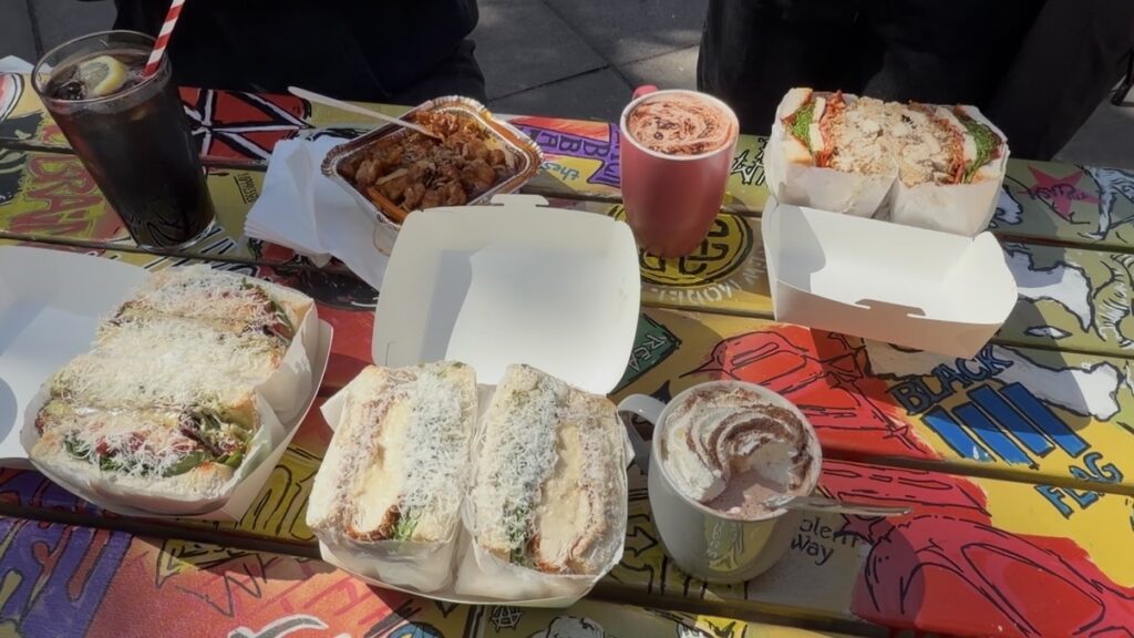 Sandwiches, a cup of coffee, a cold drink with a straw, pasta, and a cup topped with cocoa are arranged on a colorful, graffiti-style outdoor table in sunlight.