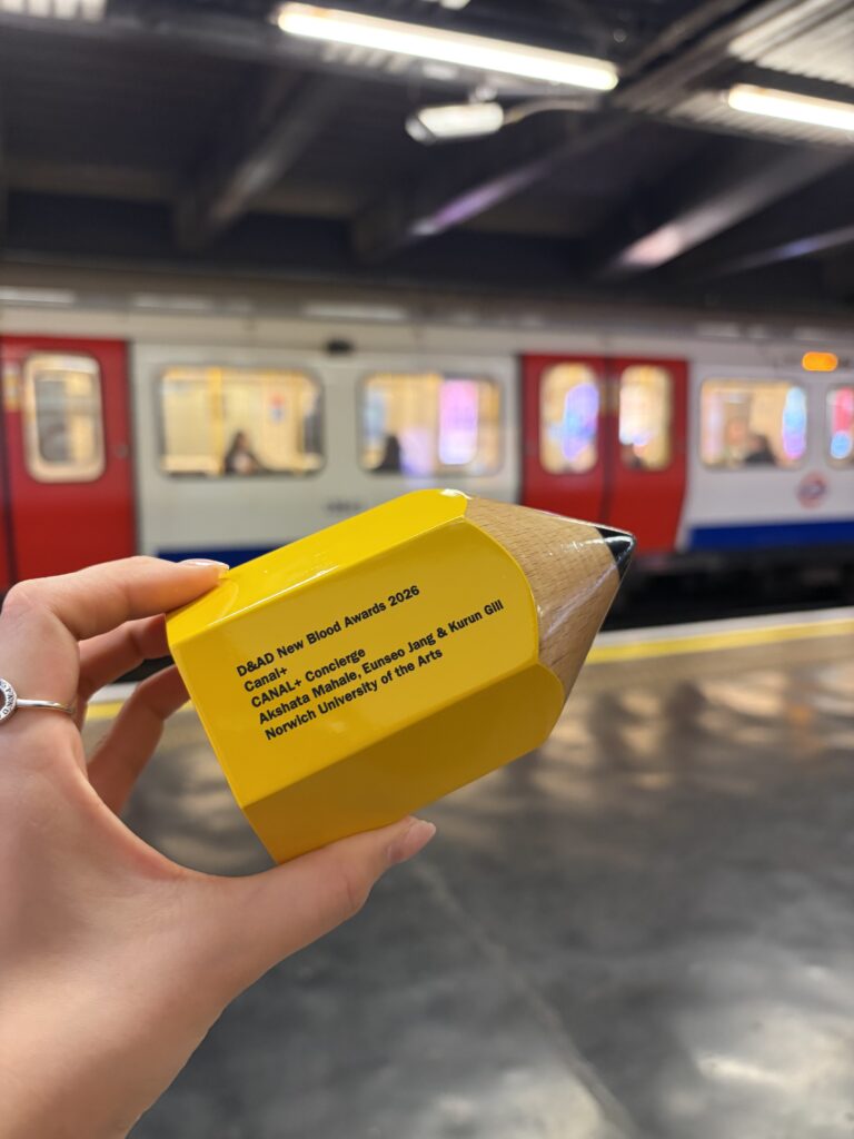 A hand holds a yellow pencil-shaped trophy with text on it at a train station platform, with a red and white train in the background.