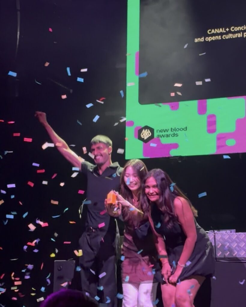 Three people celebrate on stage under falling confetti, smiling and holding a yellow award in front of a screen displaying “D&AD new blood awards.”.