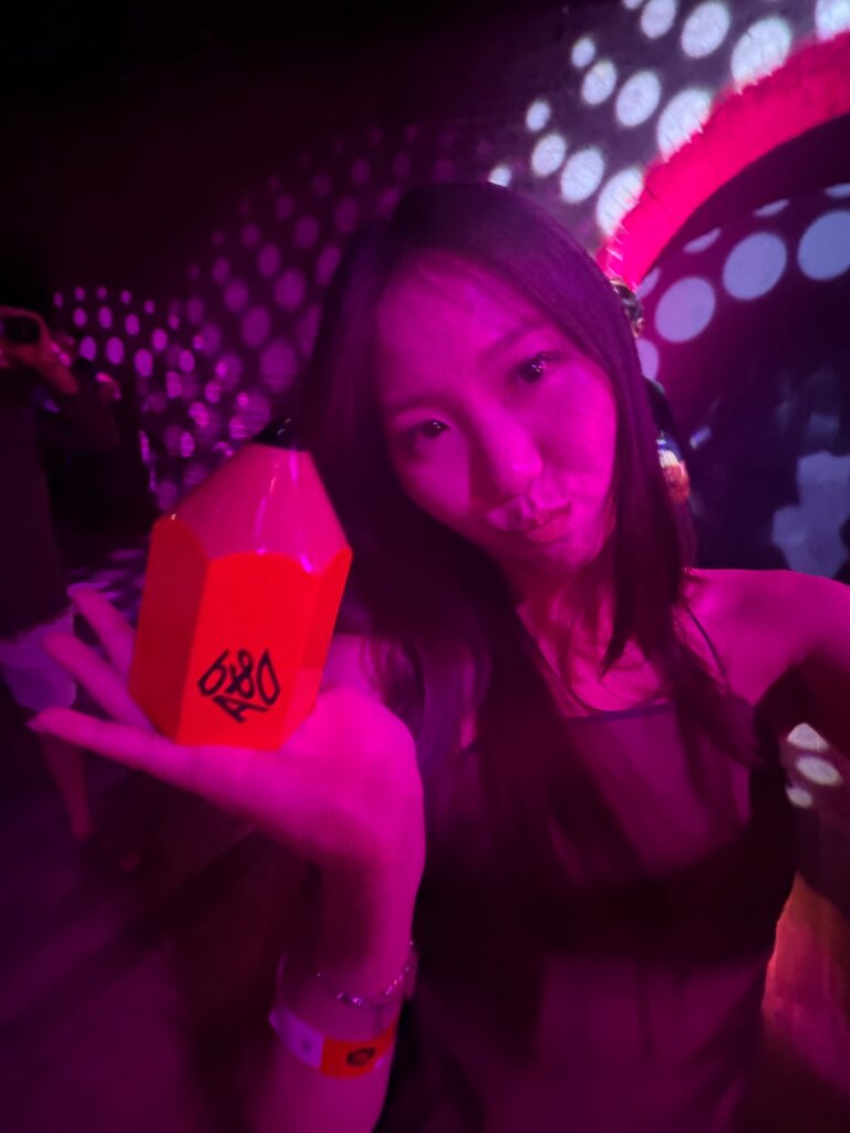 A woman poses in purple lighting, holding a red geometric object with “0690” printed on it. She stands in front of a wall with circular light patterns.