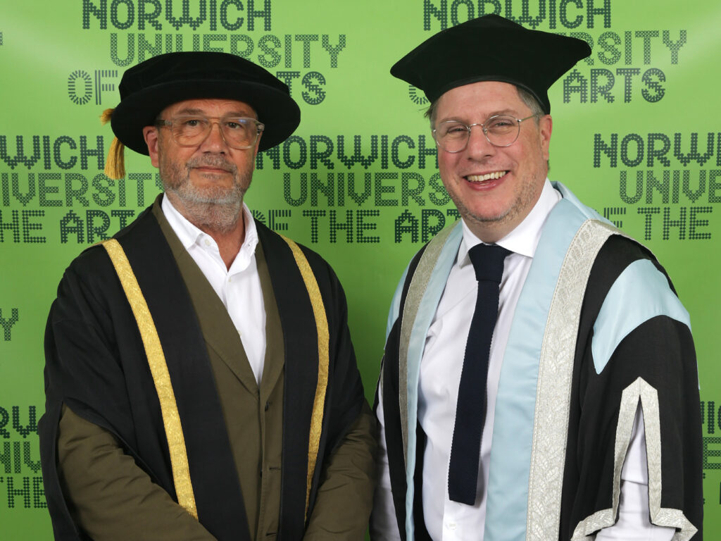 Two men in graduation academic regalia stand in front of a green backdrop with “Norwich University of the Arts” repeated in black text.