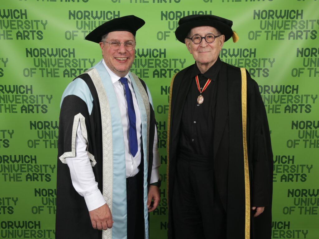 Two people in graduation academic robes and hats stand smiling in front of a green backdrop with the words “Norwich University of the Arts” repeated in the background.