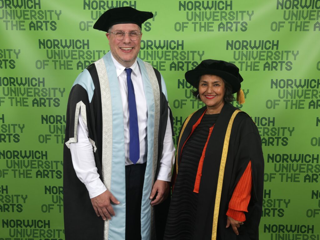 Two people in graduation academic regalia stand smiling in front of a green backdrop with “Norwich University of the Arts” printed in repeat on it.