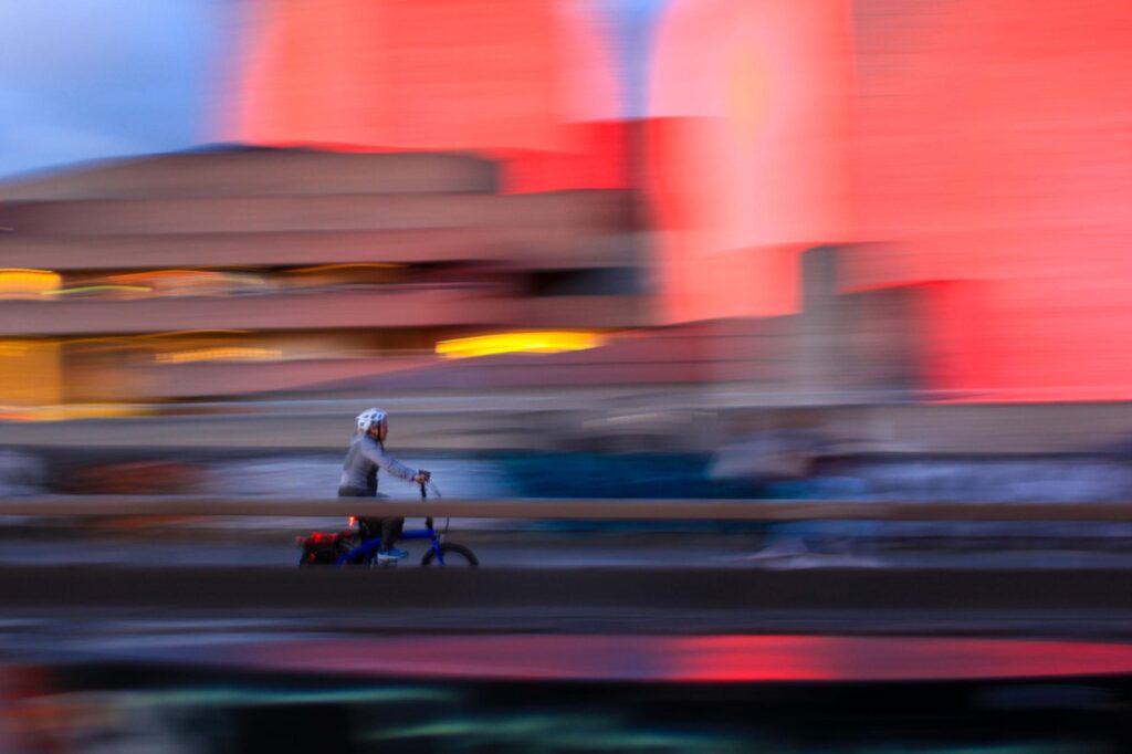 A person wearing a helmet rides a bicycle past blurred buildings with bright pink and orange lights, creating a sense of motion.