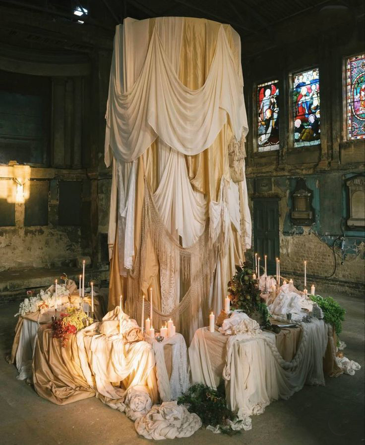 A large draped fabric installation stands in an old, dimly lit room with stained glass windows. Candles, greenery, and flowers decorate the fabric-covered tables around the centerpiece.