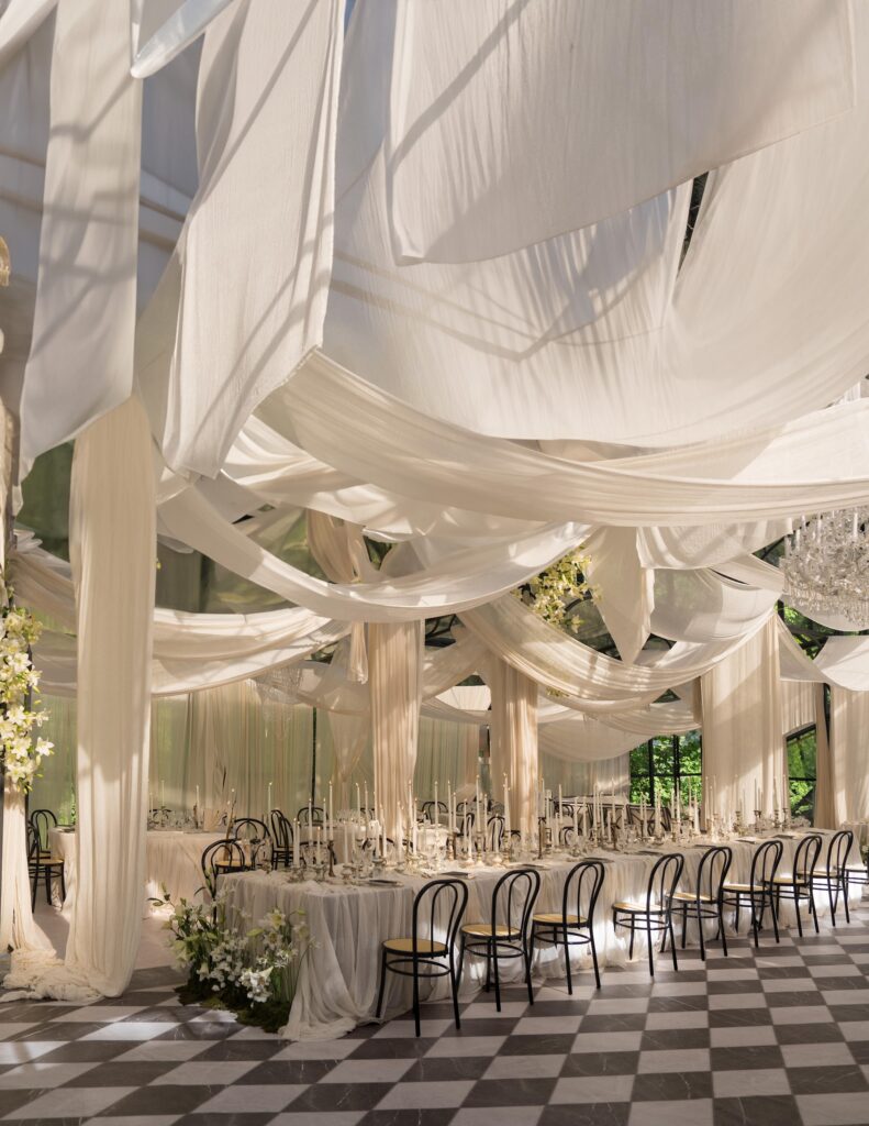 Elegant banquet hall with black chairs, white draped fabric on the ceiling, chandeliers, and long, decorated tables set for a formal event on a black and white checkered floor.