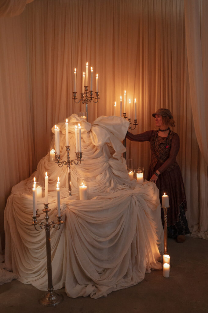 A person in vintage clothing stands beside a draped white fabric structure adorned with lit candles in ornate candelabras. The scene is softly lit, creating a warm, atmospheric, and theatrical effect.