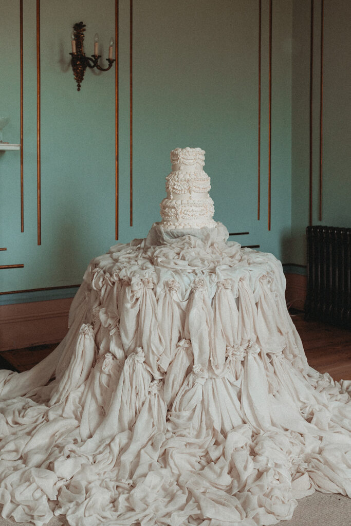 A white tiered cake sits on a table covered with flowing, draped white fabric, creating a dramatic, cascading effect in an elegant room with blue walls and gold trim.