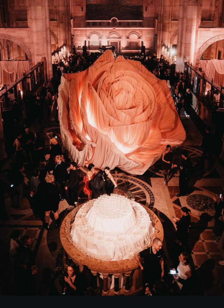A large rose-shaped installation dominates a grand hall filled with people in evening attire, with a round table of glasses in the foreground. The space is warmly lit, creating an elegant, festive atmosphere.