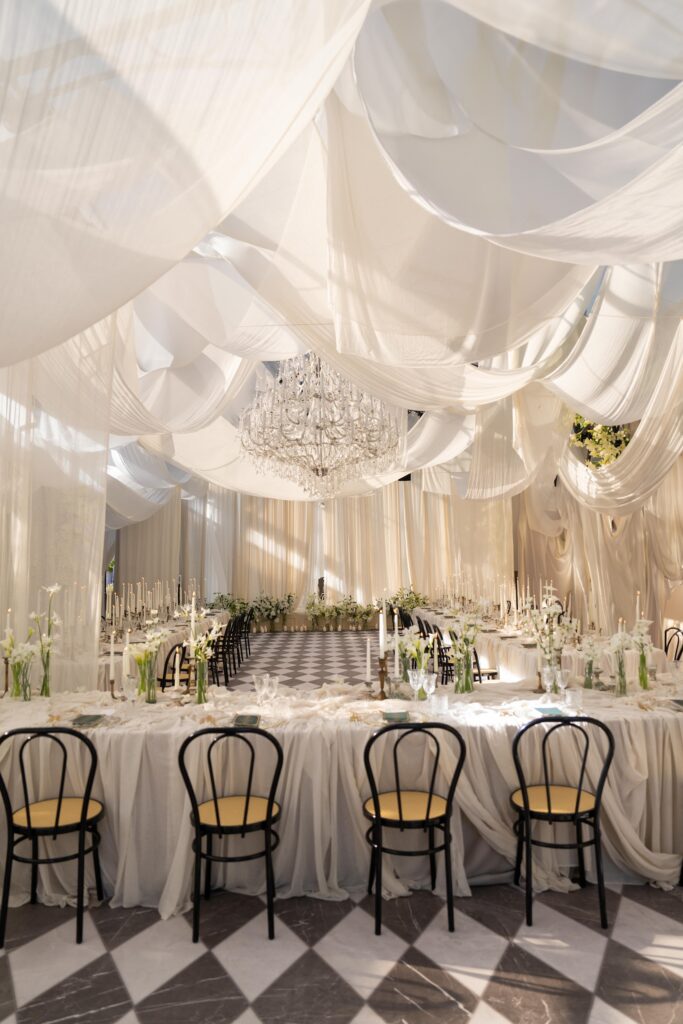 Elegant wedding reception with draped white fabric ceiling, chandelier, long tables set with white linens, flowers, candles, black chairs, and checkered floor. Natural light filters through, creating a soft, romantic atmosphere.