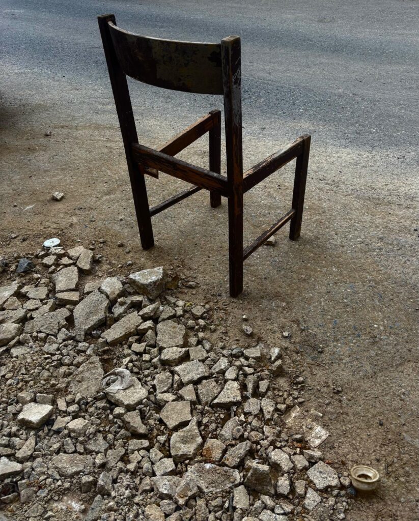 A worn wooden chair sits on bare ground next to a pile of broken concrete pieces, facing a paved road.