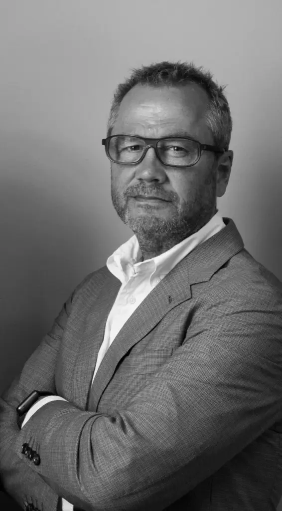 A man with short hair and glasses, wearing a suit jacket and white shirt, poses with arms crossed against a plain background. The photo is in black and white.