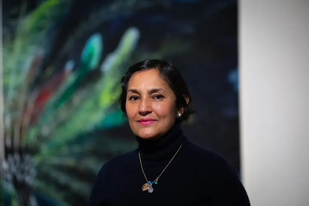 A woman with short dark hair and a dark turtleneck sweater stands in front of a colourful, abstract, blurred painting.
