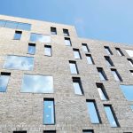 photo looking up at NUA All Saints Green building shows grey brick building with several stories of windows reflecting the blue clear sky in the background