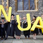 Students in black graduation gowns stand in front of a large flint brick building and holding chest high large polystyrene letters and smiling at the camera