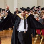 Photo of a student in graduation robes and cap with a white shirt lifting hands above his shoulders and pointing with fingers off camera with many students and attendees in the background