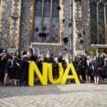 Photo of students in graduation robes throwing hats into the air to celebrate while standing next to chest high yellow letters N U A with a large flint brick church in the background with tall glass windows
