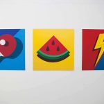 Photo of mounted graphic work shows vibrant colour prints of table tennis paddle with white circle on dark and light blue split background, a watermelon slice against a yellow background and a lighting bolt dissecting a red and blue background