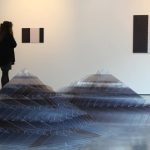 photo of the East Gallery at NUA shows a pyramid-like shape in the foreground constructed from small clear plastic sheets with various black and white pieces are on the walls and someone inspecting the work