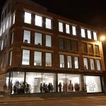 exterior photo taken at night of The East Gallery at NUA shows Cavendish House with several brightly lit rooms and a gathering of people in the ground floor of the building at an exhibit