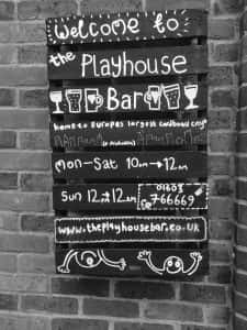 photo of a menu board on a brick wall for The Playhouse Bar in Norwich with opening times and a website URL and simple drawings of glasses and a smiling person doodle