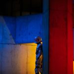 Image depicts a man in a space suit standing in a concrete scene with red, blue and yellow lights projected onto the space