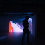 Atmospheric photo of a man walking through a dark room in front of a screen with a colourful film on at the Norwich University of the Arts MA Degree Shows