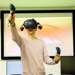 A man using the VR Lab at Norwich University of the Arts, wearing a VR headset lifting his arms up in front of a projector