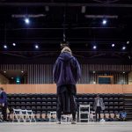A student in baggy clothes — a baggy hoody, over a long baggy skirt — faces an empty seating area from the centre of a stage, under spotlights on a high ceiling.