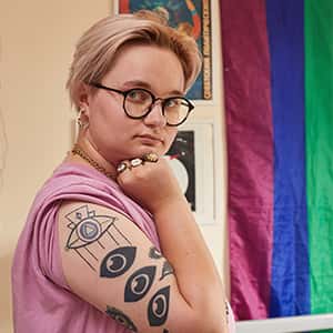 Person standing with tattoos on their arm, rainbow flag in background and they have blonde hair and glasses on