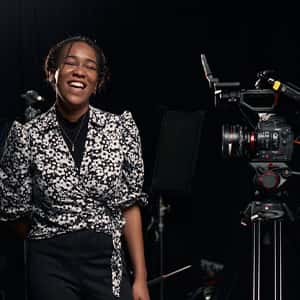Tanicha wears a black and white speckled blouse with puff shoulders, over a black top and black trousers. She is laughing, standing behind a film camera on a tripod