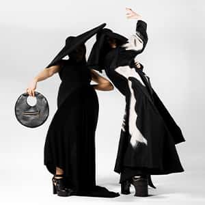 Two figures strike a pose in long monochrome dresses, with their faces obscured by the shadows from their extremely wide-brimmed hats. Together, their silhouette is almost a circle on the white background