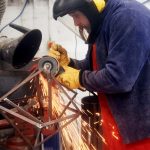 An NUA Fine Art student with full-face visor and leather protective gear grinds down welds on a geometric metal sculpture in the 3D workshop, creating bright orange sparks