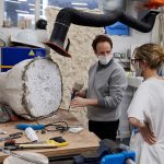 A technician in the 3D studio at Norwich University of the Arts talks to a student about their project. A large polystyrene mould, encased in concrete or clay, sits on the wooden work surface which is littered with handtools.