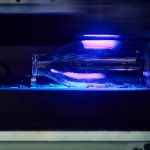 A clear glass bottle reflects vibrant violet light while being printed upon in the UV printer at Norwich University of the Arts