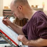 A mature student studying architecture at Norwich University of the Arts works intently on a technical drawing on one of the drawing tables in the architecture and interior design studio space.