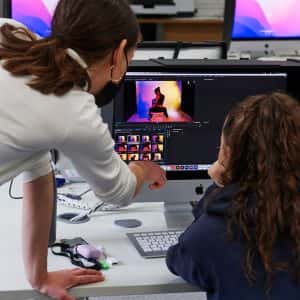 A member of staff gives 1 to 1 support to a student in the Digital Darkroom, who works on their images of a model sat facing away from the camera, with vibrant purple and yellow lighting. The student is working on a large iMac screen which has a black hood to protect the colour integrity seen.