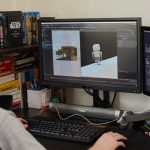 A student working with two screens in their accommodation, the desk is dark wood and has a bookshelf beside it, with books stored in stacks as well as vertical.