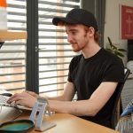 A student working at a laptop in their accommodation. The laptop is on an aluminium stand, in the background there is a plant and a TV on a bookshelf