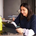 A student working with a stylus on a tablet device at a desk in their accommodation, the desk is a dark grey wood and also has a laptop and books on it