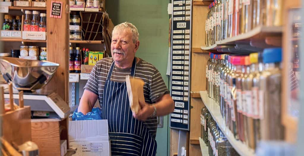 A man wearing a t-shirt and a striped navy apron is holding a paper bag and looking warmly into the camera. He is surrounded by racks of herbs and spices.