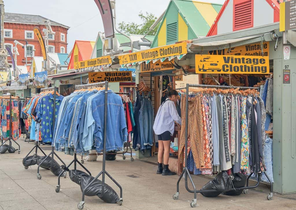 Photo of Taxi Vintage Clothing on Norwich market. Several racks of clothes stand outside a market stall with a pitched, striped roof.