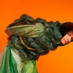 Model leaning over in a green patchwork outfit against an orange backdrop.