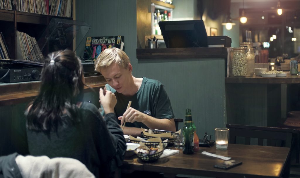 Two people eating with chopsticks at a dark wooden table. The walls are painted a dark green, and there is warm, orange lighting.