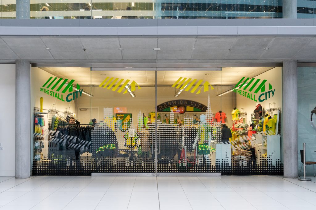 A photo of the shop front of 'On The Stall City' in the Forum, Norwich. The shopfront is made up of a large glass window and door. A Norwich city building skyline design runs across the glass taking up the whole shopfront.